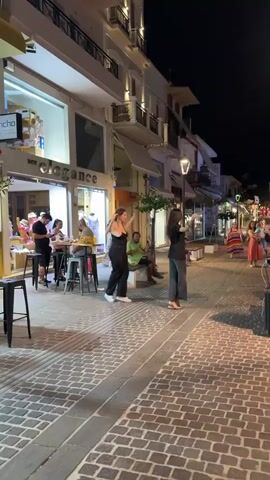 The video depicts a nighttime scene on a cobblestone street lined with businesses. On the left, an open-air restaurant or cafe area shows several patrons seated at tables and standing. A man in a black shirt and shorts stands near the tables, and other figures are visible further in the background. In the center of the street, two women are dancing. One woman, wearing a black top and pants, is in motion, her arms raised. The other, in a dark top and gray pants, is also dancing. Behind them, several people are seated on a bench, and further down the street, more individuals are gathered, some appearing to be watching the dancers. A yellow balloon is visible floating in the upper right corner of the frame. The street is illuminated by streetlights and the lights from the businesses. The buildings appear to be multi-story structures with awnings. The overall atmosphere is lively and social.