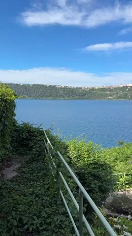 The video shows a scenic view of a blue lake under a bright sky. In the distance, a city skyline is visible along the opposite shore. A white railing is in the foreground, surrounded by lush green vegetation. A small section of a stone pathway is visible alongside a well-manicured lawn and small garden bed of flowers.