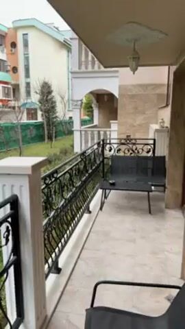 The video shows a small balcony with ornate black wrought iron railings and white pillars. A bench and chair sit on the tiled balcony. The view from the balcony looks out onto a grassy lawn area lined with green privacy netting along a fence. A few bare trees and some evergreen shrubs are scattered across the lawn.  A multi-story building is visible in the background, with a pastel color scheme and multiple air conditioning units mounted on its exterior. The sky appears overcast.