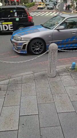 A modified, silver four-door Nissan Skyline R34 drives past on a street in Tokyo, Japan. The car has blue racing stripes and other modifications. The camera follows the car as it passes. People and other vehicles are visible on the street in the background. A DIESEL store is visible in the background. The sidewalk is paved with gray stone. A chain links concrete posts along the edge of the sidewalk.