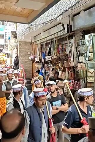 This video captures a vibrant procession moving through a narrow, bustling market street, likely in Kasol, India. A large group of men, predominantly adults and some elders, form the main part of the procession. Many are dressed in traditional Himachali caps, some with visible text like "जय शिव शंभू" (Jai Shiv Shambhu), and a mix of traditional kurtas, vests, and modern clothing. Some participants in the foreground hold staffs or ceremonial maces, one with a red sash. In the distance, a small, ornate palanquin or chariot, possibly carrying a deity, is visible, being carried by other members of the procession.

The street is lined with market stalls on the right, displaying a variety of goods such as handbags, sling bags, and other accessories. The buildings are multi-storied, featuring brickwork, exposed electrical wiring, and some security bars on windows. The architecture and traditional attire suggest a Himalayan town setting. The overall atmosphere appears festive and ceremonial, possibly indicating a religious or cultural event. The audible sound of drum beats further enhances this impression. The time of day appears to be daytime, with natural light illuminating the scene.