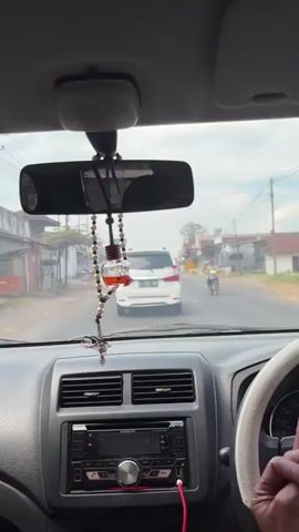 The video provides an interior view from a car driving on a road. In the foreground, the dashboard is visible with an audio player and air vents. A hand is seen on the steering wheel. A beaded car air freshener with a cross hangs from the rearview mirror. In the middle ground, a white SUV is traveling ahead. To the right of the road, a person on a motorcycle is also visible. The background depicts a semi-rural environment with buildings and vegetation along the roadside. The sky is overcast.