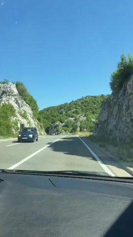 The video, filmed from inside a moving vehicle, shows a winding road surrounded by green, rocky hills under a clear blue sky. On the left side of the road, a silver car drives past. On the right side of the road, a yellow sign and a red stop sign are visible. The car drives along the road, and the scenery gradually changes as the car moves forward. The video ends as the car continues to drive along the road.