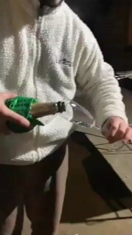 Here's a description of the video:

A person in a white fleece jacket and brown pants is pouring champagne into a flute glass. The bottle is wrapped in green fabric with gold sequins. They're outside on a patio, and the light is somewhat dim. The jacket has "LA Angels" printed on the chest. The camera is focused on the act of pouring the champagne, capturing the bubbles forming in the glass.
