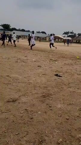A group of young men are playing soccer on a dirt field. The sky is overcast, suggesting it might be a cloudy day. The field is predominantly dirt with patches of dry grass and scattered debris. In the background, there are several low-rise buildings, possibly part of a school or residential area, with palm trees visible in the distance. The players are dressed in athletic wear, with some wearing white jerseys and shorts, and others in dark jerseys and shorts. One player in a dark jersey is actively kicking a red soccer ball. The overall scene depicts a casual soccer match or practice session taking place in an outdoor, somewhat undeveloped setting. There is no visible text in the image.