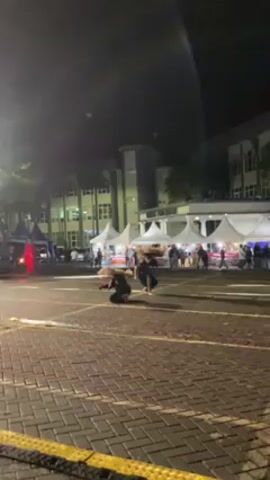 Here's a concise description of the video:

The video captures a performance involving two dancers in traditional attire at night. The dancers, wearing elaborate headdresses, are performing on a brick-paved parking lot. In the background, there are several white tents and a multi-story building. There are people milling around the tents, suggesting some kind of event or market. There is a person in a red costume on the left side. The lighting is dim.