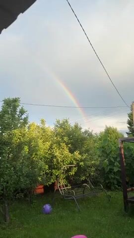 A rainbow arcs across a partly cloudy sky, visible above lush green trees. Power lines crisscross the upper portion of the frame. In the foreground, the top of a metal swing set is visible. The camera is positioned at a low angle, looking up at the sky.