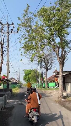 Two people ride a scooter down a paved road. The person in front is wearing a brown shirt and the person behind is wearing a yellow shirt. They are both facing away from the camera. The person behind is carrying a white cylindrical object. There are trees and power lines on either side of the road. Buildings with red roofs are visible in the background. The sky is blue and clear.