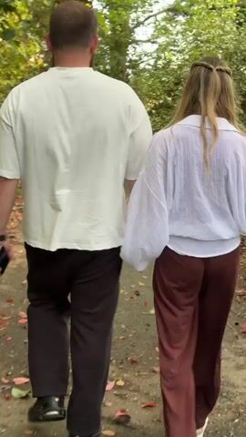 A couple, a man and a woman, are walking hand-in-hand down a tree-lined path. The man is wearing a white t-shirt, dark brown pants, and black shoes. He has short hair and a beard and has sunglasses perched on the back of his head. The woman has long blond hair with a styled braid and is wearing a white blouse with long sleeves and dark brown pants. They are walking away from the camera through a green wooded area with leaves scattered on the path.
