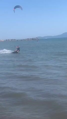 A person is kiteboarding on a body of water under a clear blue sky. The kiteboarder is wearing a dark t-shirt and dark shorts and is holding onto a kiteboarding bar. The kiteboarder is in motion, creating a spray of water behind them as they ride on a kiteboard. The water is choppy with moderate waves. In the background, there is a distant shoreline with hills and mountains, and several other kiteboarders or sailboats can be seen on the water. The overall scene depicts a sunny day with active water sports.