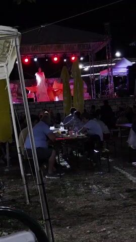 A nighttime outdoor event is captured, with a stage set up in the background. On the stage, a performer in a white dress with voluminous sleeves is dancing, illuminated by red spotlights. Behind the performer, a backdrop displays images. A banner above the stage reads "OPEN FESTIVAL 'NOVA VODA' VENETA". In the foreground, people are seated at tables, some eating and drinking, while others are watching the performance. The environment appears to be a grassy area with some tents and umbrellas providing shade.