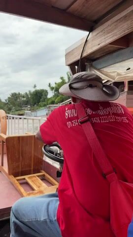 The video begins with a close-up shot of the back of a person wearing a red t-shirt, a wide-brimmed hat, and blue jeans, sitting at the helm of a boat. The t-shirt has white text on it, likely in a foreign language, and a red strap goes across the person's chest. The person is holding a steering wheel, and the boat appears to be moving forward on a river. Wooden chairs are arranged on the deck of the boat. There is a life preserver ring on the left side of the boat, near the front. The riverbanks are lined with trees and some buildings. The sky is overcast. The camera pans to the left, revealing more of the boat's deck and the river ahead. Several other boats are visible in the distance along the river. The video ends as the boat continues to travel down the river.