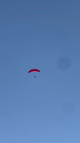 A person is paragliding against a clear blue sky. The paraglider's canopy is red, and the person is a dark silhouette suspended beneath it. The paraglider appears to be descending or gliding through the air. There are no other objects or individuals visible in the frame. The environment is simply the sky, with some circular smudges on the lens.