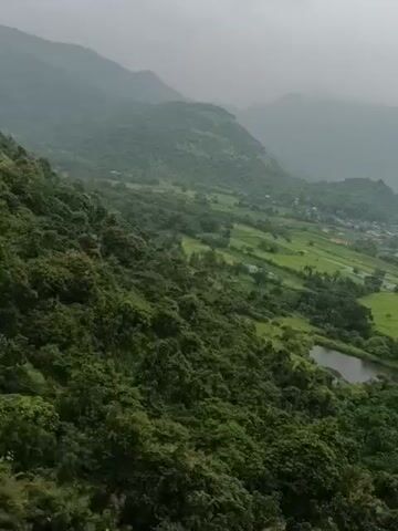 The video displays a wide shot of a lush green valley from a high vantage point. The valley is filled with trees, agricultural fields, and a small village. In the foreground, dense green foliage covers a steep slope. As the camera pans, it reveals rolling hills and mountains in the background, shrouded in a light mist. A small body of water, possibly a pond or lake, is visible in the middle ground. The overall impression is of a serene and natural landscape. The video appears to be a stabilized drone shot, with a slight tilt to the right.