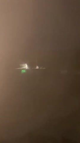 The video shows an airplane on a foggy tarmac at night. The airplane is illuminated by lights, including a green light on its fuselage. The visibility is extremely poor due to the fog, and distant lights can be seen through the fog, suggesting the presence of a town or other structures. The video is shot from a static position, possibly from a window.