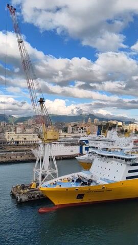 The video shows a large yellow ship, "MEGA EXPRESS THREE", docked in a harbor with a city visible in the background. A large crane stands beside the ship. The sky is blue with numerous puffy white clouds. The shot is taken from an elevated position, likely from another ship or high vantage point. The video is oriented vertically.
