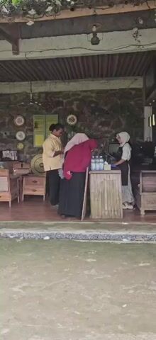 The video shows a group of people standing at a counter made of bamboo. To the left, a person wearing a yellow shirt and dark pants is looking down at their phone. In the center, two individuals wearing hijabs are bending over the counter, appearing to be selecting or purchasing items. One person is wearing a pink hijab and a red outer garment, while the other is wearing a dark hijab and a black skirt. On the right, another person wearing a white hijab and an apron is standing behind the counter, possibly serving or assisting the customers. There are several bottles of water and a small television set on the counter. The background features a stone wall with decorative plates and a wooden structure. The ground in the foreground appears to be covered with small stones or pebbles.