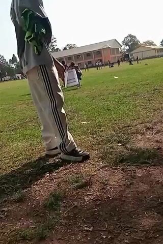 The video shows a grassy field with several people in the background, likely students. There are buildings in the distance, possibly a school. The foreground is dominated by the lower half of a person's body, who is wearing white pants with black stripes and dark sneakers. The person is standing on a dirt and grass area. The sky is visible and appears to be clear. There is some debris scattered on the grass. The video is shaky and the camera is pointing towards the field.