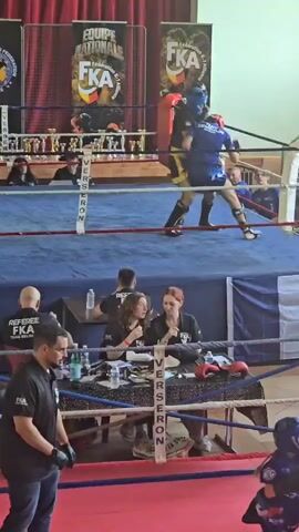 The video depicts two individuals engaged in a kickboxing match within a boxing ring. One participant is wearing a blue uniform and headgear, while the other is in a yellow and black uniform with headgear. The fight is occurring in front of a banner that reads "Equipe Nationale FKA" and "Fédération de Kick Boxing," indicating it is a sanctioned event. Referees and judges are visible around the ring, monitoring the fight and scoring. One man in a black shirt that says "REFEREE FKA" is walking around the outside of the ring as the fighters compete. A table with water bottles and other items is positioned ringside, where officials appear to be taking notes. The environment suggests the event is taking place indoors in a hall or gymnasium-like setting.