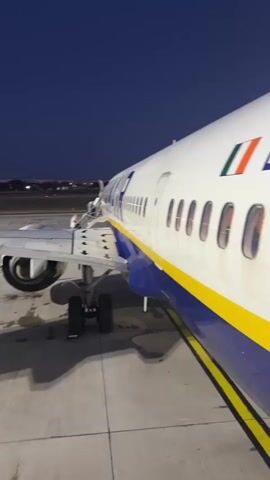A Ryanair airplane sits on a concrete apron at dusk. The plane is white with a yellow stripe and blue lower body. The landing gear is visible below the wing. A boarding ramp leads up to the airplane's entrance. In the background, lights from a distant building are visible against the dark blue sky.