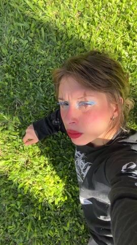 A woman with heavy blue eyeshadow, dark red lipstick, and blush, wearing a black long-sleeved shirt with a white graphic, lies on a patch of green grass, looking up at the camera. She raises her right hand to her forehead, shielding her eyes slightly. Her hair is light brown and appears wet or damp. The grass is well-lit and vibrant. The camera appears to be held above the woman, looking down at her.