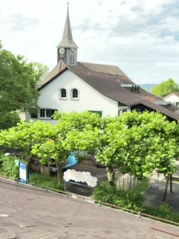 The video shows a view of a white church with a steepled roof and a clock tower. The church is surrounded by lush green trees, and the sky above is partly cloudy. The camera zooms in slightly on the church, revealing architectural details such as arched windows and a pointed spire. There are no people or discernible actions visible in the video. The environment appears to be outdoors on a bright day, likely in a town or village setting.