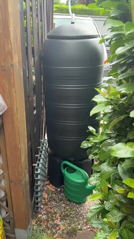 A close-up shot focuses on a tall, dark green water butt positioned next to a wooden fence. To the left of the water butt, a green plastic watering can rests on the ground. The ground is covered with small stones and fallen leaves. Lush green foliage is visible to the right of the water butt, partially obscuring the view. The video appears to be shot outdoors, possibly in a garden or backyard, with the lighting suggesting it is daytime.