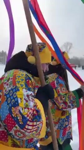 The video shows a group of people on snow, holding a pole structure with colorful ribbons attached. The ribbons are waving in the wind. People are dressed in colorful clothes. It appears to be a winter celebration or festival. A few trees are visible in the background.