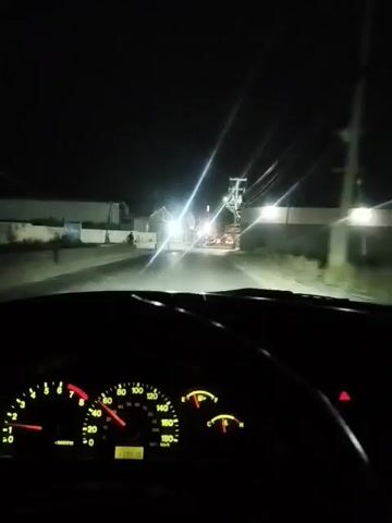 A car is moving down a street at night.  Streetlights and lights from buildings illuminate the area.  Power lines are visible overhead. The car's dashboard is visible, displaying various gauges.  The speedometer indicates the car is traveling approximately 30 kilometers per hour.  In the distance, it appears there are people standing on the side of the road.  The video appears to be shot from the driver's perspective.
