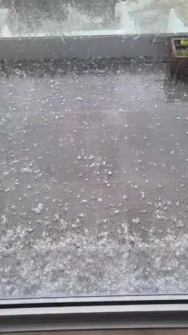 The video shows a ground-level view from indoors looking through a window during a hailstorm. The ground is covered in hail, with a higher concentration of hail closer to the window. Rain is also visible on the window, and the outside view is blurred. The window frame is visible at the bottom of the frame. The hail appears to be small and round. Beyond the window, blurry greenery can be seen.