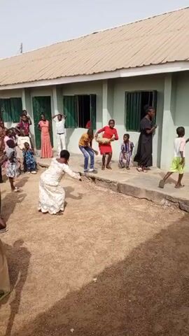 A group of children and adults are gathered in front of a building with green doors and window shutters. The building appears to be a school or community center.  A woman in a white dress with floral patterns is dancing in the foreground, holding a red object in her hand.  Behind her, several children are dancing on a concrete area in front of the building.  A group of people are standing in front of the building, observing.  Some faces can be seen looking out the windows.  The ground is dirt.  The weather appears sunny.