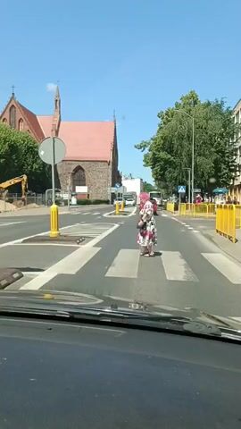 This is a video clip taken from inside a vehicle, looking out at a street scene. The primary subject is a woman with bright pink hair, wearing a floral dress and carrying a handbag, who is crossing a pedestrian street. In the background, a large, red-roofed church with a tall, dark spire dominates the skyline. Adjacent to the church, construction work is visible, with an excavator present. The sky is clear and blue, suggesting a sunny day.

The street itself has clear white pedestrian crossings. Several cars are visible on the road, and some yellow barriers are placed along the roadside, likely indicating a temporary obstruction or construction area. A traffic sign with a blue circular background and a white arrow pointing to the left is also visible. The overall setting appears to be an urban or town environment, with the church suggesting a historical or central location. The bright sunlight and clear sky indicate it is daytime, likely midday or early afternoon. There is no discernible text in the video. The activity is straightforward: the woman is crossing the street while vehicles navigate the road.