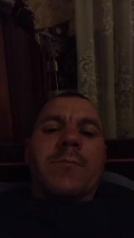 A close-up, selfie-style video shows a bald white man with a mustache and light skin. He appears to be indoors in a dimly lit room. He is wearing a dark shirt. His facial expression changes from neutral to grimacing as he speaks. The background is blurred, but hints of furniture and curtains are visible.