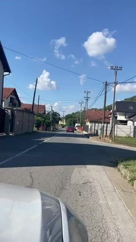 A static point-of-view camera captures a daytime scene from the perspective of a vehicle moving forward on an asphalt road. The sky above is clear blue with sparse white clouds. Utility poles and power lines extend along the right side of the road, which also features a white solid line on the shoulder and a broken white line in the middle. Buildings with red roofs and fences line both sides of the road. A red car is visible in the distance, driving away from the camera. A white van is parked on the right side of the road. The video concludes with the vehicle continuing to drive forward.