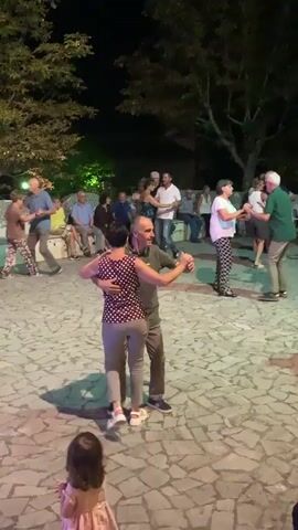 The video shows a nighttime outdoor gathering with several couples dancing in a paved area. The ground is covered in irregularly shaped, light-colored stones, with some sections illuminated by purple and green lights. Trees with dark foliage are visible in the background, and a darker night sky is overhead. In the foreground, a young child with long brown hair and wearing a light-colored top is seated, facing away from the camera.

Several people are dancing in pairs. The most prominent couple in the center of the frame consists of a man with short brown hair wearing a dark green shirt and grey pants, and a woman with short dark hair wearing a patterned top and grey pants. They are dancing in a close embrace. To their right, an older couple is dancing; the man has white hair and wears a light blue shirt and blue jeans, while the woman wears a pink top and dark pants. Further to the right, another couple is dancing, with the man wearing a dark shirt and the woman wearing a white top and patterned pants.

In the background, more people are visible, some dancing and others sitting on a curved bench or standing around. There's a general atmosphere of a social event or celebration. The lighting suggests it's evening or night, with artificial lights creating colored accents on the ground. The overall scene depicts a lively, communal dancing activity in an outdoor setting.