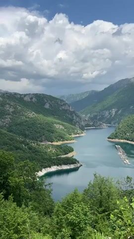 The video shows a scenic view of a long, winding river nestled between lush green mountains under a cloudy sky. The river is calm and blue, reflecting the sky.  A series of floating platforms is visible in the middle of the river. The surrounding mountains are covered in dense forest. The sky is a mix of blue and white, with fluffy clouds. The foreground is slightly obstructed by trees and vegetation.