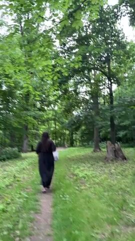 A person wearing a long black dress walks away from the camera down a dirt path through a wooded area. The person is carrying a white object in their left hand. The path is surrounded by lush green grass and trees with full canopies. The lighting suggests it is daytime.