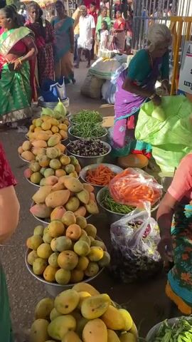 This video shows a busy outdoor market scene. In the foreground, there are several large metal bowls filled with fruits and vegetables. One bowl contains light green gourds, another contains a pile of mangoes, a third holds smaller yellow fruits, a fourth has dark purple eggplants, and another contains bright orange carrots. Green beans are also visible in a bowl. A woman in a purple and green outfit is visible on the right, handling produce. Behind the stalls, a crowd of people are walking through the market. Some individuals are wearing traditional Indian clothing. Various stalls and awnings are set up, and bags of goods are stacked behind the produce displays. The ground appears to be dirt or gravel. The overall impression is a vibrant and active marketplace.