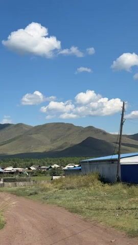 A dirt road leads into a small village nestled at the base of rolling green hills under a bright blue sky with fluffy white clouds. Several houses with visible roofs are scattered throughout the village, some with fences and trees nearby. The foreground consists of dry grass and a wooden fence. The overall scene depicts a rural, natural landscape.