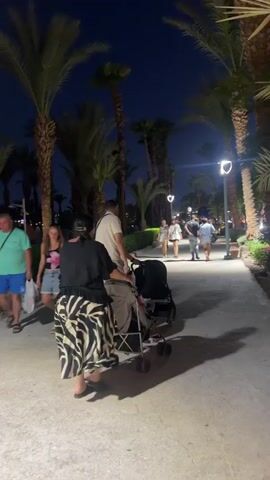 The video shows a nighttime scene on a paved walkway lined with palm trees and illuminated by decorative lamps. The walkway appears to be part of a resort or park area. In the foreground, a person is pushing a stroller from behind, with another person walking beside them. The person pushing the stroller is wearing a black top and a black and white patterned skirt. The person walking beside them is wearing a light-colored shirt and shorts. Further down the walkway, several other people are walking in the same direction. Two women in the mid-ground are walking together, one wearing a pink and white floral top and denim shorts, and the other wearing a white top and denim shorts. Another couple is visible further ahead, along with other individuals strolling along the path. The overall atmosphere is relaxed, with people enjoying an evening walk in a tropical setting.