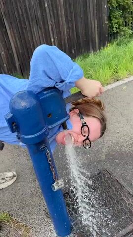 A woman in a blue shirt and beige pants is standing by a blue water pump outdoors. She bends over and drinks from the pump, then straightens up, spits out some water, and laughs. She walks away, wiping her face. The background includes a wooden fence, green vegetation, a utility pole, and a house.