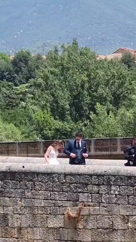 A wedding party walks across a stone bridge surrounded by lush green trees. In the foreground, a bride in a white wedding dress is followed by a groom in a dark suit. To their right, another man in a dark suit walks ahead, holding a bottle of water. The background shows a hillside with houses and a clear sky.