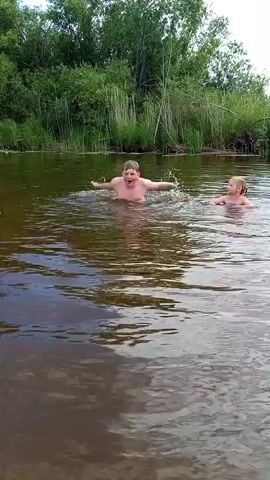 In this video, two children are playing in a body of water, likely a river, with a backdrop of lush green trees and reeds. The water is murky brown and disturbed by the children's movements, creating ripples and splashes.

In the first few seconds, one child is in the center of the frame, arms raised, as they make a significant splash in the water, appearing to be jumping or diving in. Another child, further to the right and slightly further away, is also in the water, their head visible above the surface.

As the video progresses, the splashes subside, and both children are seen swimming or wading in the water. The child who was initially in the center is now to the left of the frame, and the other child is to the right. Both appear to be enjoying themselves, with their faces visible and their movements suggesting playful activity. The overall mood seems to be one of fun and recreation on a warm day.

The natural setting suggests an outdoor activity, possibly during daylight hours given the illumination of the scene. The presence of dense vegetation along the bank indicates a rural or natural environment. No text is visible in the video.
