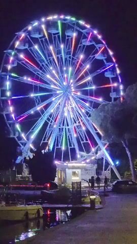 A Ferris wheel is lit with colorful lights at night. The wheel is spinning and the lights are changing colors. There are people standing around the Ferris wheel, and there are some boats in the water in the foreground. The scene is dark, and the Ferris wheel is the brightest object in the frame.