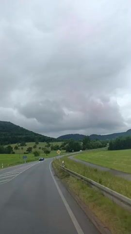 This media file captures a view from a vehicle traveling on a winding road through a rural, hilly landscape. The sky is overcast with dramatic, dark clouds, suggesting a potentially stormy or moody atmosphere. The road is paved and appears to be a two-lane highway with a white line marking the center. A guardrail runs along the right side of the road, separating it from a grassy embankment and fields.

The landscape is characterized by rolling green hills covered in trees and meadows. Scattered trees dot the hillsides, and in the distance, taller trees form a dense forest along the crests of the hills. There are no visible people in the immediate foreground, but a dark-colored car is seen in the distance on the left side of the road, indicating ongoing traffic. A yellow diamond-shaped road sign with a black border is visible on the right side of the road, likely indicating a hazard or change in road conditions. White and black marker posts are also placed along the roadside.

The overall impression is one of a scenic drive through the German countryside under a heavy, cloudy sky. The time of day is difficult to pinpoint precisely due to the overcast conditions, but it appears to be daytime. The weather contributes to a sense of drama and a somewhat subdued, perhaps melancholic, mood. The location cues, such as the road signs and the typical European rural landscape, are consistent with the provided context of Reutlingen, Germany.