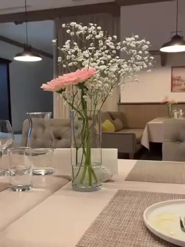 A clear, medium shot captures a table setting in a restaurant or dining area. The table is covered with a beige tablecloth and patterned placemats. A clear glass vase, holding two pink gerbera daisies and several sprigs of baby's breath, sits in the center of the table. A glass carafe and several drinking glasses are also visible. The remains of a meal are visible on a white plate near a fork. In the background, upholstered seating, wooden paneling, and other tables can be seen. The lighting is soft and warm, with hanging pendant lights illuminating the scene. The camera moves slightly closer to the flower arrangement.