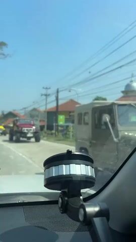 The video appears to be captured from inside a vehicle, looking out through the windshield. The foreground shows a car accessory attached to the windshield, possibly a phone mount. The road ahead is visible, with several vehicles. A red off-road vehicle with large tires is in the center of the frame, followed by a yellow Jeep and a tan pickup truck. A yellow van is also seen further down the road. There are houses and buildings lining the road, with power lines overhead. The sky is clear and blue, indicating a sunny day. There are trees and foliage on the sides of the road. The video shows movement, suggesting the vehicle is in motion.
