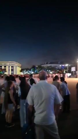 This media file captures a nighttime scene in Baikonur, Kazakhstan, likely an outdoor event or gathering. A large crowd of people is visible, appearing to be milling about in an open area. In the background, a brightly lit, modern building with multiple stories stands prominently, suggesting it might be a cultural center, administrative building, or a commercial establishment. Trees are scattered around the building and the open space. The sky is a deep twilight blue, indicating that it is either early evening or late evening. The lighting from the building and some distant lights creates a festive or illuminated atmosphere. People are dressed in casual attire. The overall mood seems relaxed and social, with individuals and groups interacting. There is no visible text in the media file.