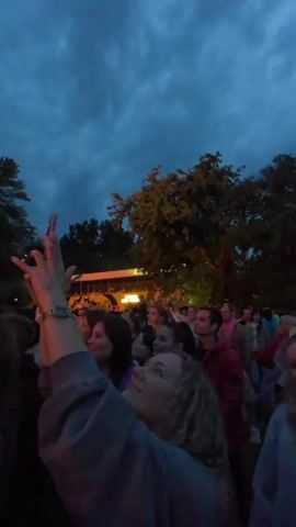 A crowd of people are gathered outdoors at dusk. The sky is dark blue with clouds. Trees are visible in the background. Some people are holding up their phones, possibly recording or taking pictures. There appears to be a stage or structure with lights in the distance. The people are facing the stage. The overall atmosphere seems to be that of an outdoor event or concert.