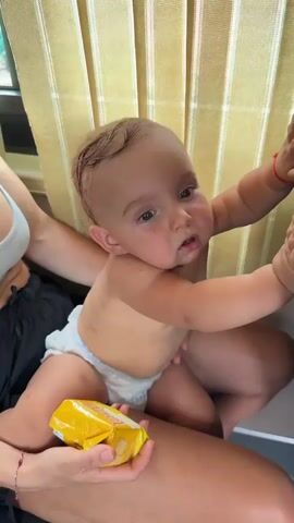 The video features a baby, likely around one year old, sitting on someone's lap. The baby is wearing a diaper and has a red bracelet on its right wrist. A yellow packet, possibly of snacks, is held in the hands of the person sitting. The background is blurred, showing vertical striped curtains or blinds, suggesting an indoor setting that might be a vehicle or a room with such window coverings. The lighting appears to be natural, indicating daytime. The baby's expression shifts from a neutral gaze to a wide smile. The person holding the baby is only partially visible, with their arms and lap in view. The overall mood is playful and heartwarming, likely a candid moment captured during a trip or outing.